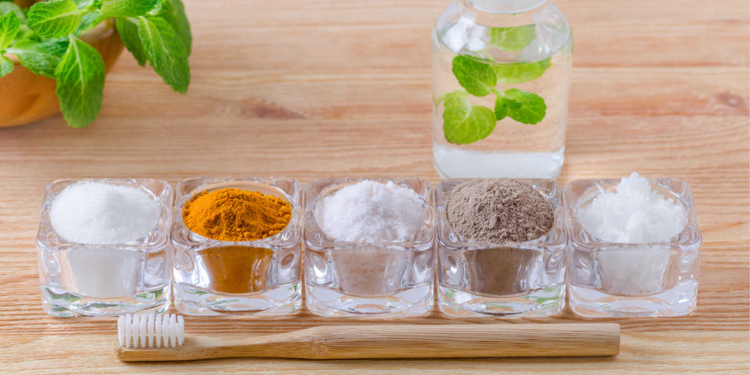 five glass containers with ingredients to make homemade mouthwash. Mint is in the background and a toothbrush is in front